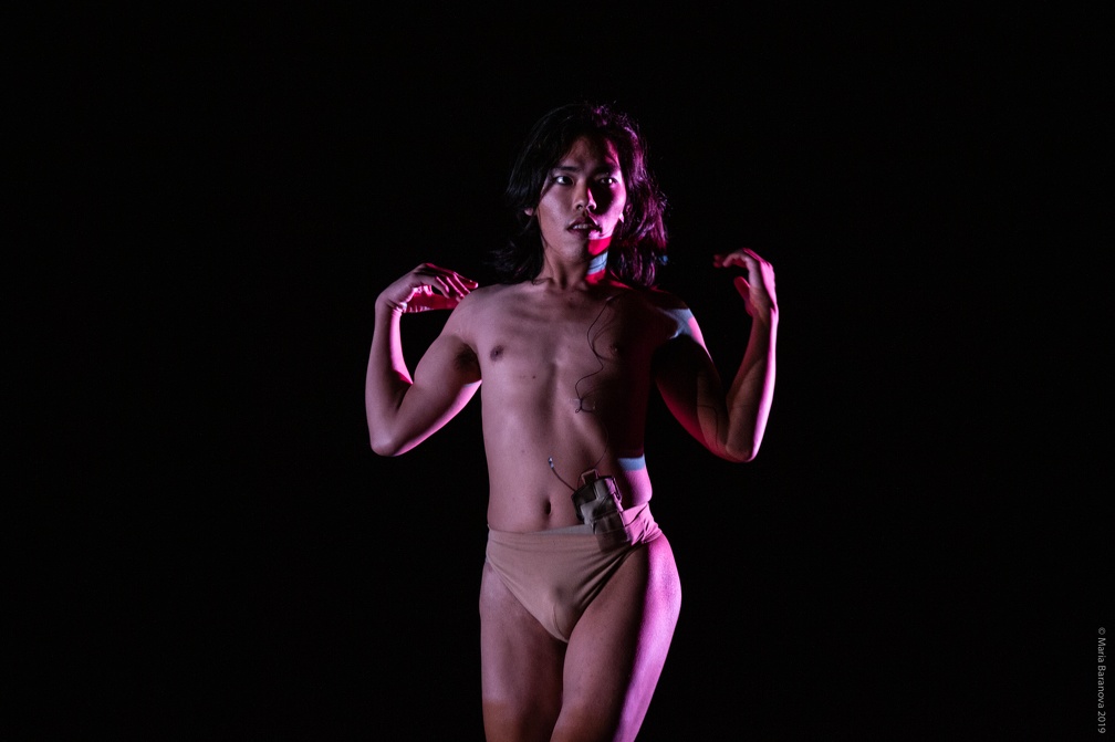 Anh Vo stands facing forward and looking slightly off into the distance wearing a beige dancer's belt with a mic pack and wire hanging from the belt to their face. Anh's hair falls gently around their face with hands raised to their shoulders. They are in soft pink and colorful light from their left hand side.