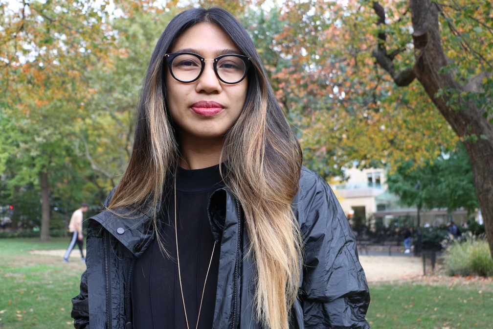 Kristel Baldoz, with trees and gras sin the background, faces forward in a black windbreaker with dark and light hair falling onto her shoulders and glasses framing her face.