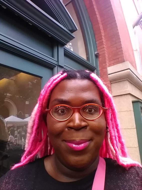 Olaiya Olayemi, outside in front of a brick building, faces the camera with a slight smile. She is in a dark knit sweater with a pink strap, pink lip stick, orange glasses, and pink braids.