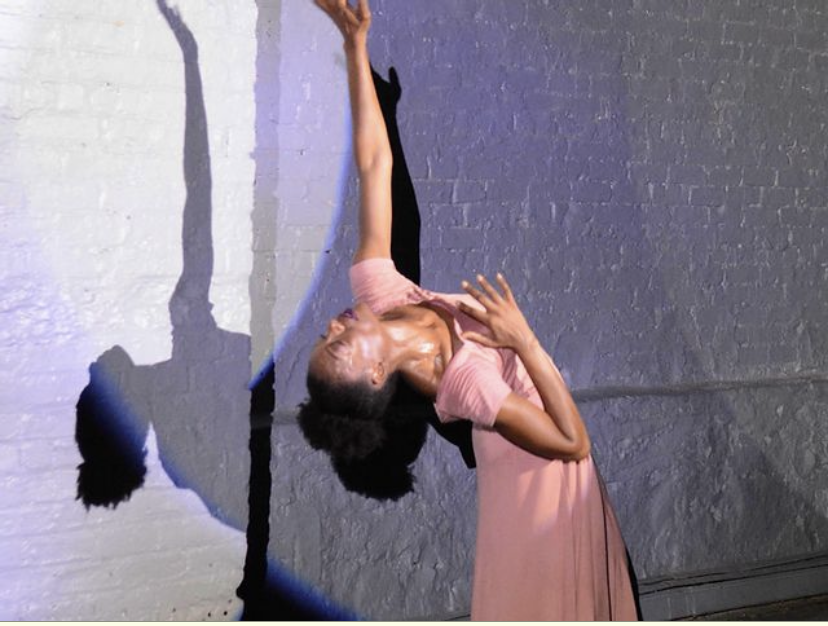 Marla Nicole Robertson bends backward in performance, face toward the ceiling, a white brick wall behind her. She is in a light pink gown with a sweetheart neckline with her hair gathered up and back atop her head.